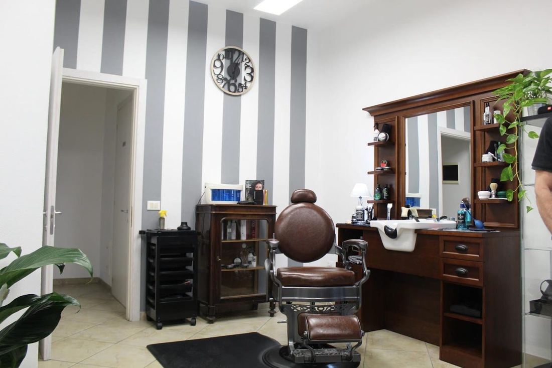 L'interno del negozio: carta da parati a righe verticali, poltrona da barbiere vintage in pelle marrone e consolle in legno scuro